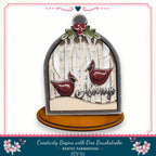 A decorative birdhouse with a snowy winter scene inside, featuring two red birds. The birdhouse is mounted on a wooden base and has a decorative border.