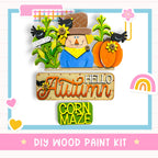 The image is a colorful and whimsical DIY wood paint kit featuring a scarecrow, pumpkins, and a corn maze, all set against a background with a rainbow and hearts.