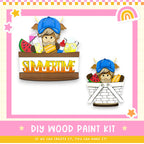 The image is a colorful illustration of a DIY wood paint kit, featuring a cartoon character wearing a blue hat and holding a basket filled with various summer-themed items such as watermelon, lemon, and a rainbow.
