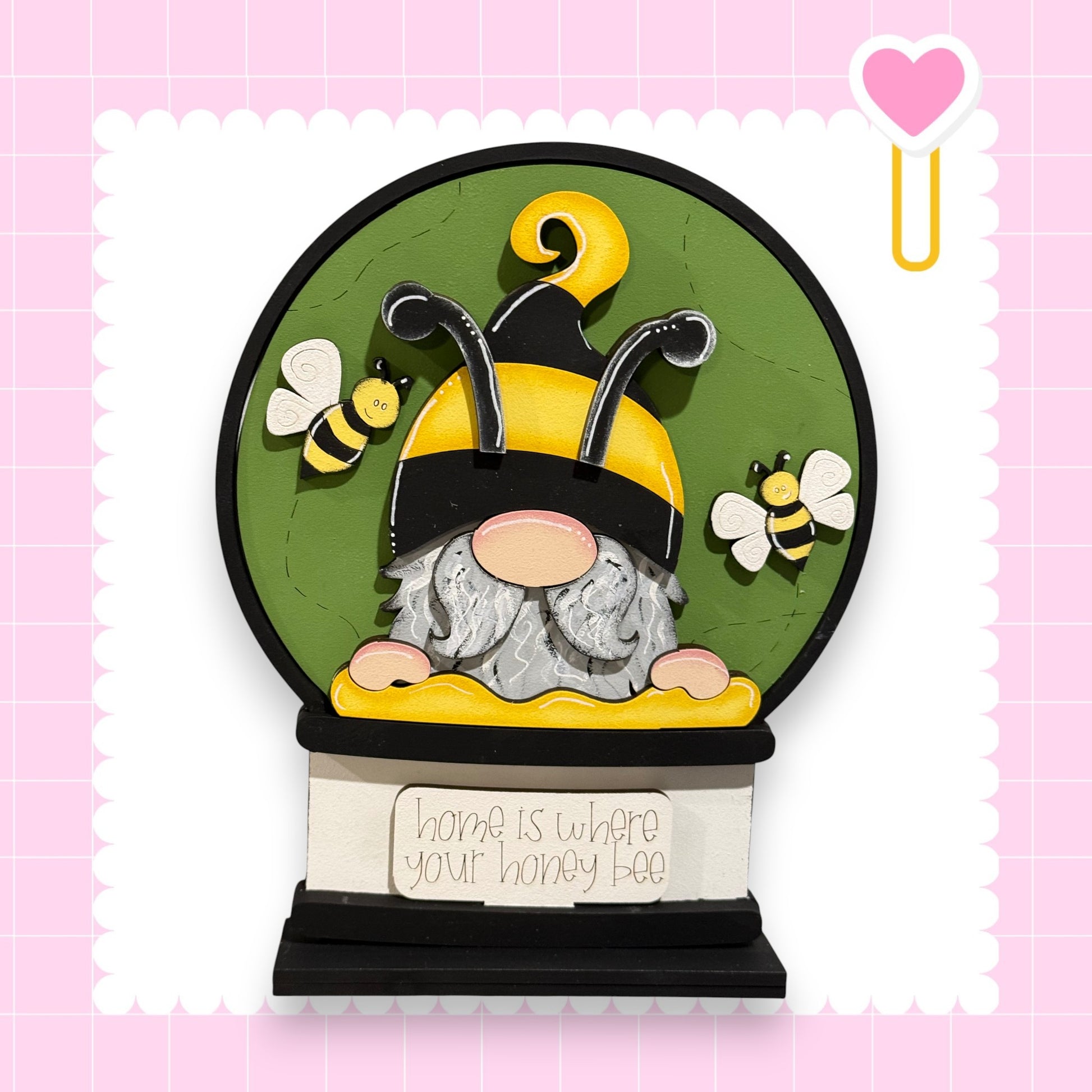 A gnome wearing a yellow and black striped hat is depicted inside a green glass globe, surrounded by two bees. The globe is set against a pink background with a scalloped edge.