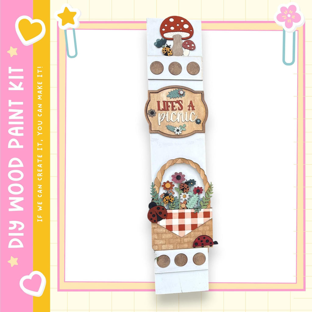 The image is a colorful and whimsical painting kit featuring a wooden frame with various decorative elements such as a mushroom, flowers, and ladybugs. The kit appears to be designed for creating a picnic scene, with the words "Lives a Picnic" and "If we can create it, we can make it" written on it.