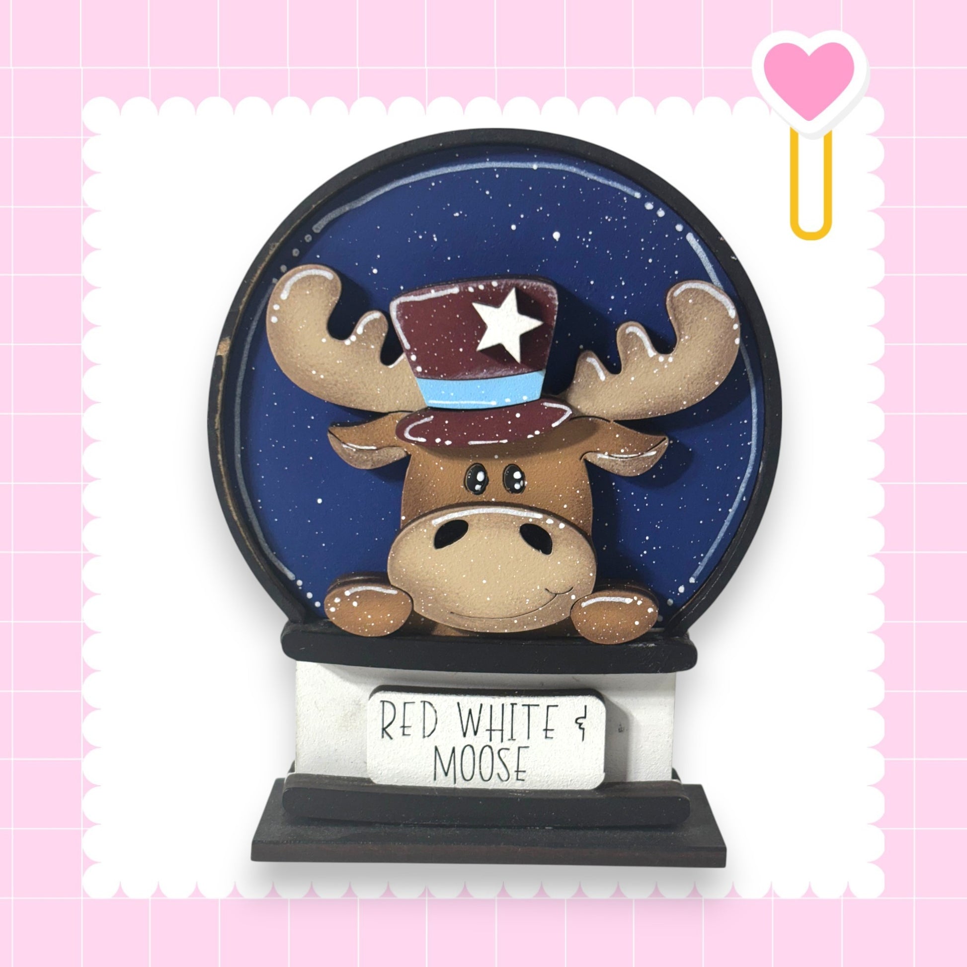A snow globe featuring a cartoon moose wearing a top hat with a star, set against a blue background with a pink heart and a white heart.
