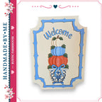 A hand-made wooden sign with a blue border and the word "Welcome" written on it. The sign features a decorative arrangement of pumpkins and a vase.
