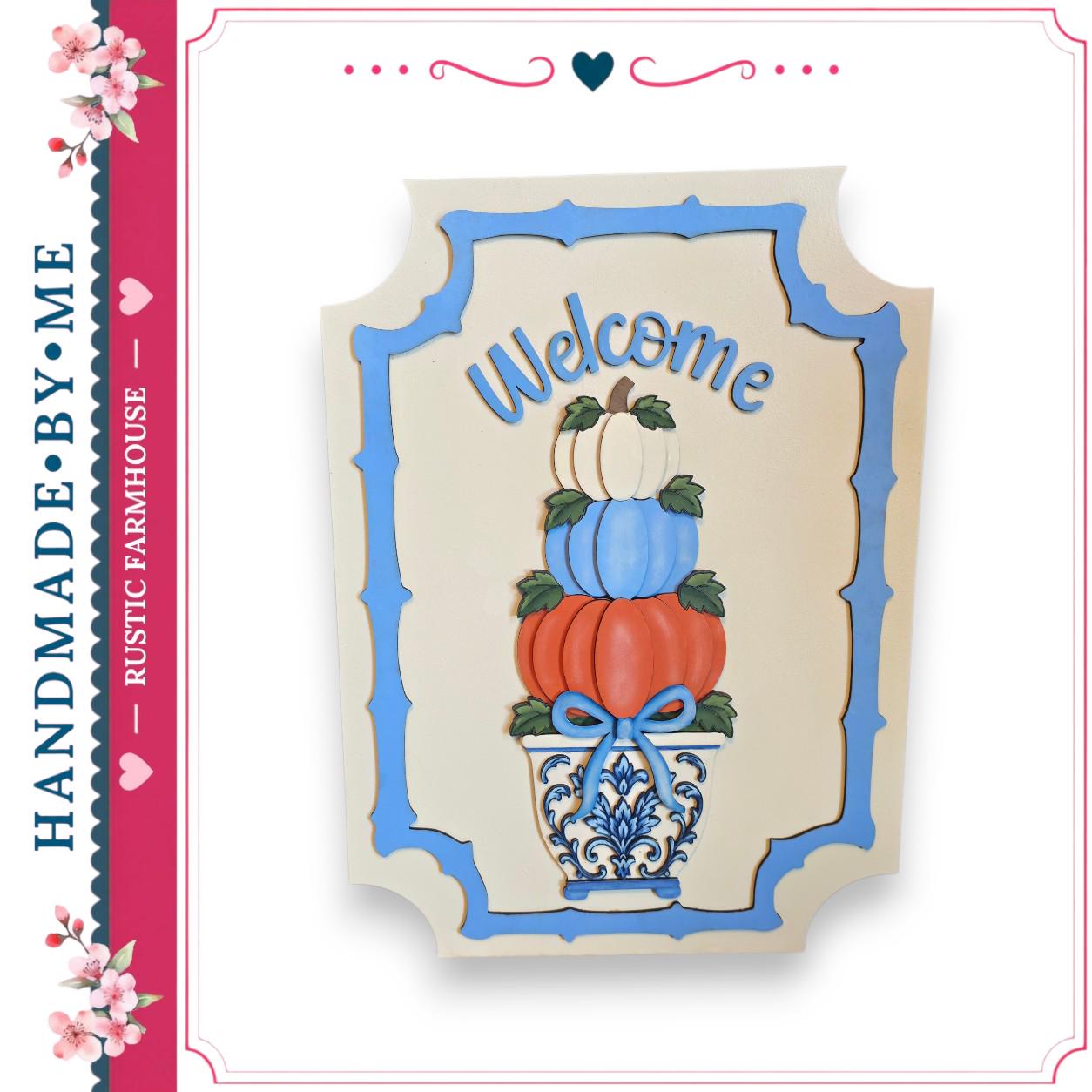 A hand-made wooden sign with a blue border and the word "Welcome" written on it. The sign features a decorative arrangement of pumpkins and a vase.