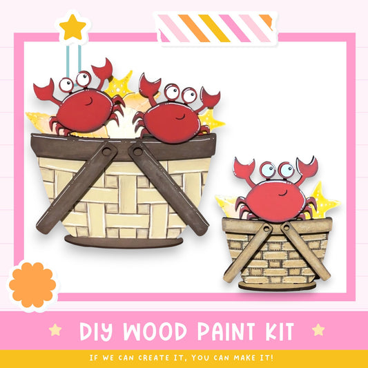 a DIY wood paint kit with a pink and white background. There are three wooden baskets, each containing a red crab. The baskets are arranged in a row, with the largest basket on the left, the medium-sized basket in the middle, and the smallest basket on the right. The crabs are sitting inside the baskets, and there are stars and a flower decoration on the left side of the image. The text at the bottom of the image reads "DIY WOOD PAINT KIT" and "IF WE CAN CREATE IT, YOU CAN MAKE IT!"