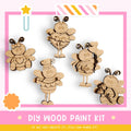 The image is a DIY wood paint kit featuring five wooden bee characters in various poses, including one holding a heart and another holding a bouquet of flowers. The kit appears to be made of wood and is designed for painting.