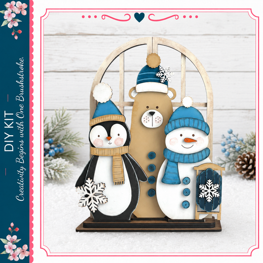 A wooden cutout of a penguin, a teddy bear, and a snowman, all wearing winter hats and scarves, is displayed on a wooden stand against a snowy background.