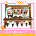 A wooden ice cream stand with various ice cream flavors displayed on cones, along with a sign that reads "ICE CREAM". The stand is made of wood and has a pink and white striped border. The image also includes a DIY wood paint kit and a statement encouraging creativity.