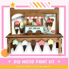 A wooden ice cream stand with various ice cream flavors displayed on cones, along with a sign that reads "ICE CREAM". The stand is made of wood and has a pink and white striped border. The image also includes a DIY wood paint kit and a statement encouraging creativity.