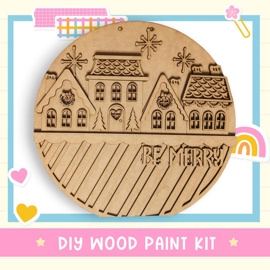 A wooden paint kit with a festive design featuring houses and snowflakes, along with the text "Be Merry" and a "DIY WOOD PAINT KIT" label.