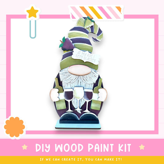a whimsical wooden gnome-like figure holding wine glasses and a sign that says "I want glasses". The background is a pink and white color scheme with a star and a flower. The image includes text that reads "DIY WOOD PAINT KIT" and "IF WE CAN CREATE IT, YOU CAN MAKE IT!"