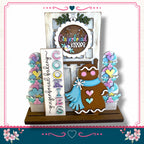 A gingerbread house-themed display with a gingerbread cookie, a sign that says "Gingerbread", and a wreath. The display is set against a white background with a pink border.