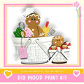 A DIY wood paint kit featuring a gingerbread man and a small child, along with various craft supplies, such as a paintbrush and a palette, all set against a pink and white background.