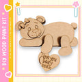 A wooden bear figure with a heart-shaped message attached to it, set against a pink and yellow background.