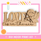 The image is a promotional poster for a DIY wood paint kit. It features a wooden sign with the word "LOVE" written on it, along with a cute cartoon character resembling a cherub or angel. The poster emphasizes that the kit allows for creativity and encourages viewers to create their own designs.