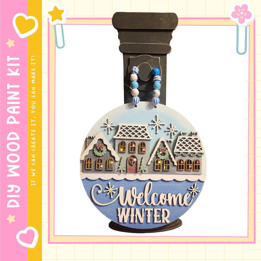 A wooden paint kit with a winter-themed design, featuring a snow globe with a village scene and the text "Welcome Winter" on a blue background.