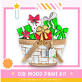 A DIY wood paint kit with a basket filled with painted wooden shapes, including bows and presents, against a pink and white background.