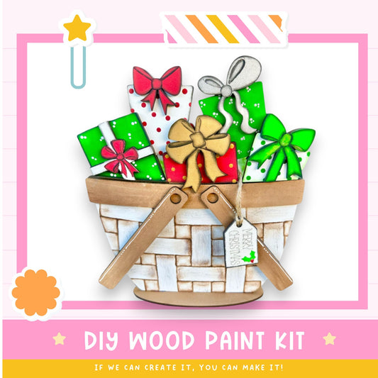 A DIY wood paint kit with a basket filled with painted wooden shapes, including bows and presents, against a pink and white background.