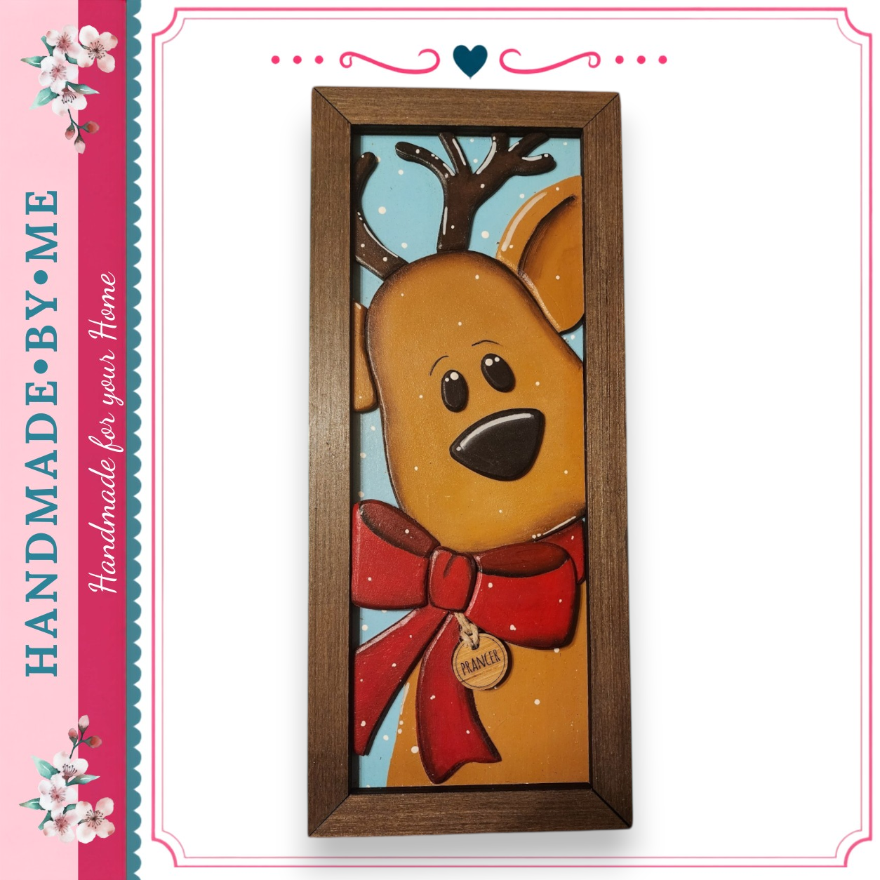 A wooden picture frame with a painted reindeer wearing a red bow and a tag that says "PANIEK". The reindeer has antlers and is looking directly at the camera.