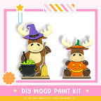 two cartoon moose characters dressed in Halloween costumes, one wearing a witch's hat and the other a carved pumpkin. The moose characters are holding various Halloween-themed items, such as a cauldron filled with green apples and a broom. The image also includes text that reads "DIY WOOD PAINT KIT" and "IF WE CAN CREATE IT, YOU CAN MAKE IT!"