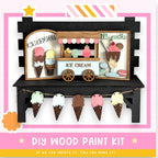 The image is a colorful and whimsical depiction of an ice cream shop, featuring a wooden ice cream stand with various ice cream flavors and toppings. The shop is decorated with paper cutouts of ice cream cones and scoops, creating a playful and inviting atmosphere. The image also includes a DIY wood paint kit, suggesting that the viewer can create their own ice cream shop scene.