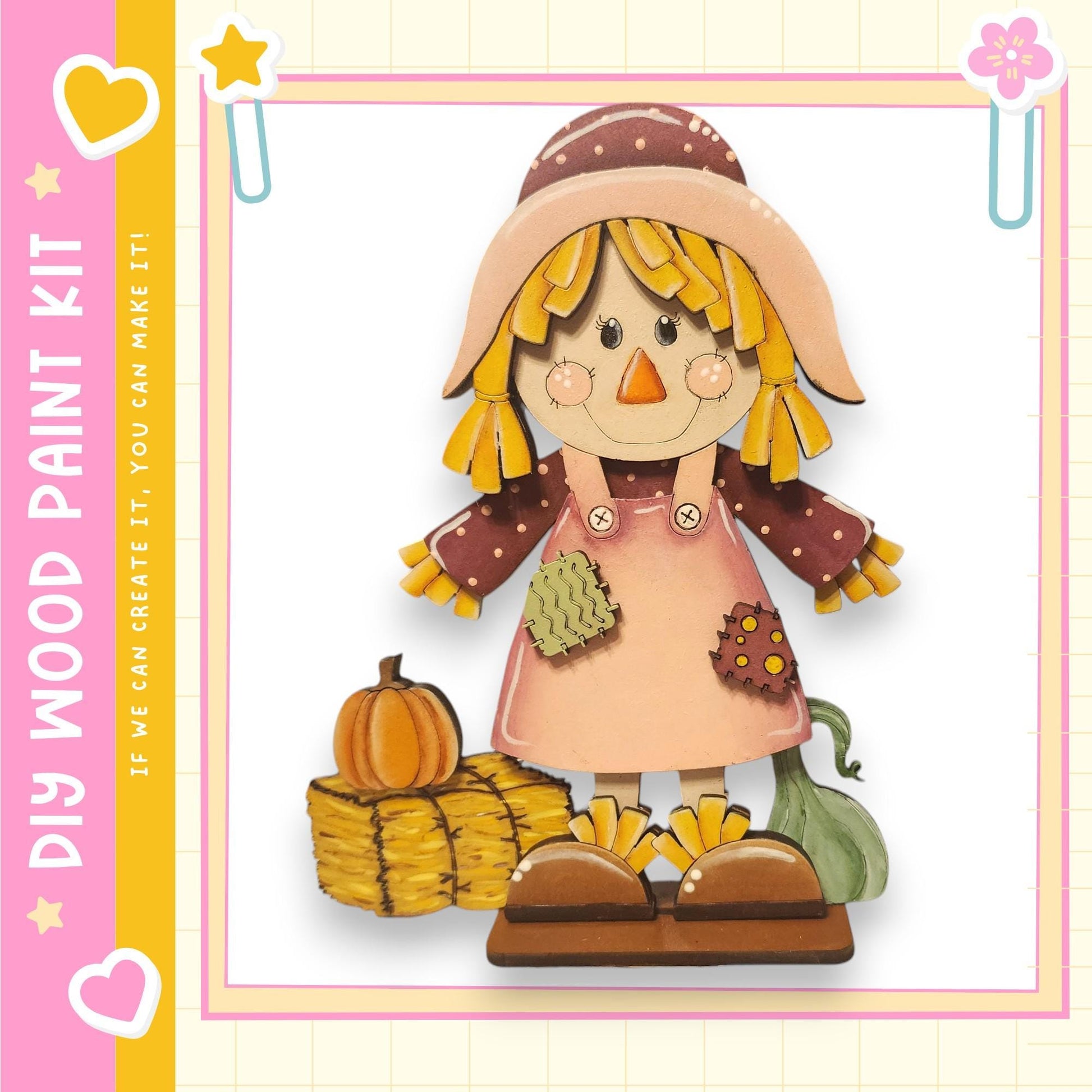 A wooden paint kit depicting a scarecrow with a pumpkin and hay bales, set against a pink and yellow background with a heart and star motif.