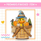 A whimsical illustration of a duck wearing a witch's hat, surrounded by pumpkins and a bird, sitting in a basket.