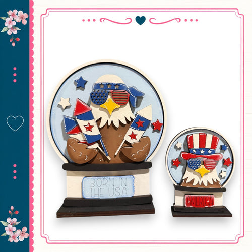 two wooden snow globes, each depicting an eagle wearing patriotic attire. The text on the snow globes reads "Born in the USA".
