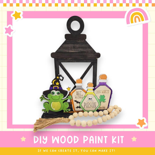 a DIY wood paint kit with a lantern, a frog, and three bottles labeled "BAD WINS POSSESSION" and "Frog Warts". The kit appears to be made of wood and includes a tassel. The image also includes a rainbow and stars, suggesting a festive or whimsical theme.