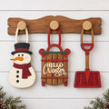 a wooden board with three wooden ornaments hanging on it, including a snowman, a sled, and a shovel.
