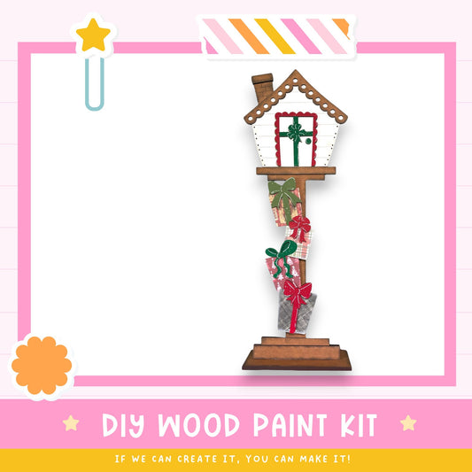a DIY wood paint kit, with a wooden house-shaped object painted in a festive style with presents and a star decoration. The kit appears to be made of wood and is designed for painting.