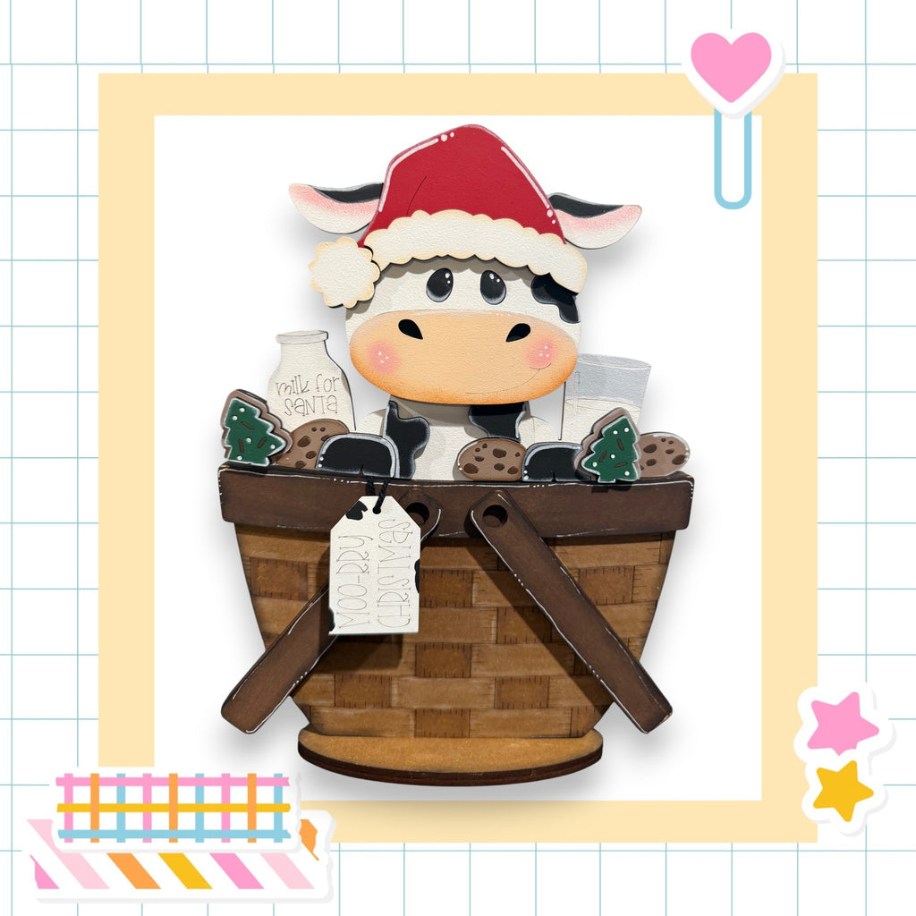 A wooden cow wearing a Santa hat sits in a basket filled with various items, including a milk carton and cookies.