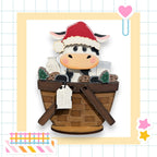 A wooden cow wearing a Santa hat sits in a basket filled with various items, including a milk carton and cookies.