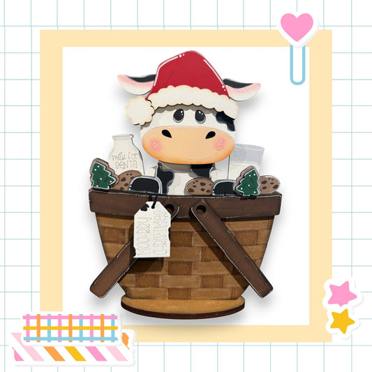 A wooden cow wearing a Santa hat sits in a basket filled with various items, including a milk carton and cookies.