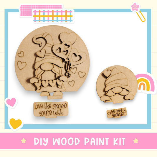 The image is a DIY wood paint kit featuring two wooden gnome-shaped figures, one larger and one smaller, with hearts around them. The kit includes a sign that says "Love the gnome you're with" and a smaller sign that says "Be my gnome".