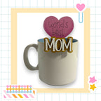 A white mug with a heart-shaped decoration on top, surrounded by various colorful and decorative elements, including a heart-shaped sticker and a pink heart-shaped object.