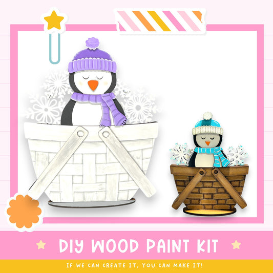 a DIY wood paint kit, with a penguin and a snowman in a basket, along with a pink and white striped border. The kit appears to be made of wood and is designed for creating custom wooden figures.
