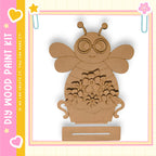 A wooden paint kit depicting a bee with flowers in a vase, set against a pink and yellow background with decorative elements.