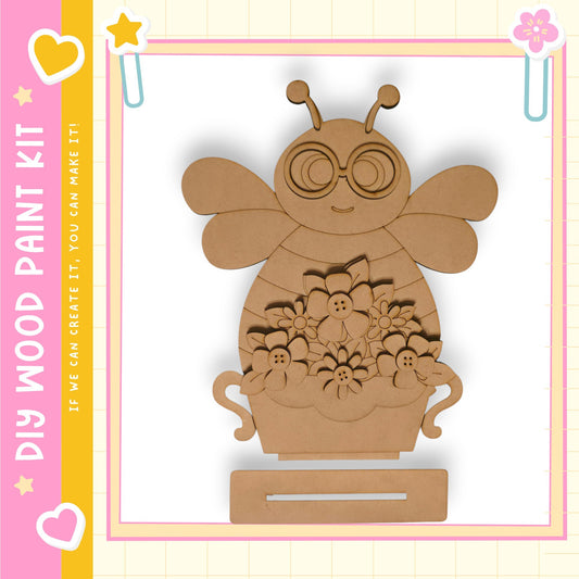 A wooden paint kit depicting a bee with flowers in a vase, set against a pink and yellow background with decorative elements.