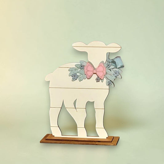 A white wooden lamb figure with a bow and floral decorations is displayed on a wooden base.