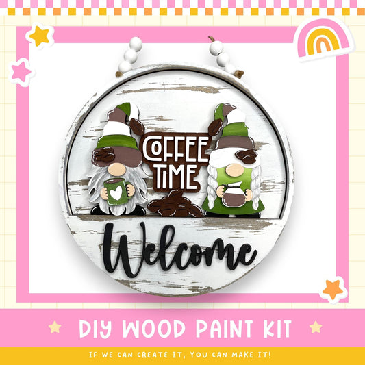 a DIY wood paint kit for creating a wooden sign with a coffee-themed design. The kit includes a wooden sign with the words "Coffee Time Welcome" and two gnome-like figures holding coffee cups. The kit is presented in a circular frame, and the text "DIY WOOD PAINT KIT" is displayed at the bottom.