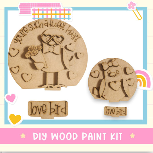 The image is a DIY wood paint kit featuring two wooden bird-shaped figures, one larger and one smaller, with hearts and a bow around them. The kit is labeled "Love Bird" and includes a sign that says "DIY WOOD PAINT KIT".