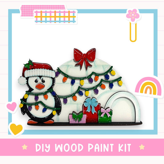 A DIY wood paint kit featuring a penguin wearing a Santa hat and surrounded by a Christmas tree with colorful lights and presents.