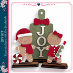 two gingerbread men, one wearing a red hat and the other wearing a red dress, standing in front of a green sign with the word "JOY" written on it.