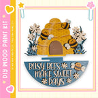 A wooden bee hive with three bees on top, surrounded by flowers and a sign that says "Busy bees make sweet days".