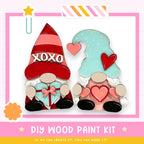 two wooden gnomes holding hearts, with one gnome wearing a red hat with the word "XOXO" and the other wearing a blue hat with a heart. The image also includes text that says "DIY WOOD PAINT KIT" and "IF WE CAN CREATE IT, YOU CAN MAKE IT!"