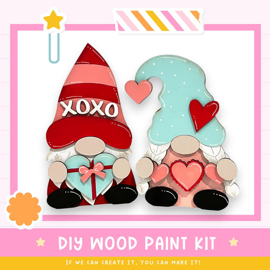 two wooden gnomes holding hearts, with one gnome wearing a red hat with the word "XOXO" and the other wearing a blue hat with a heart. The image also includes text that says "DIY WOOD PAINT KIT" and "IF WE CAN CREATE IT, YOU CAN MAKE IT!"