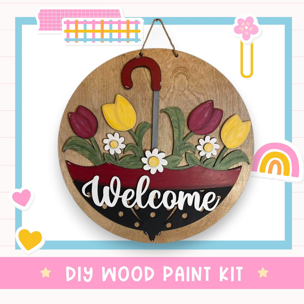 A wooden sign with a red umbrella handle and flowers, displaying the word "Welcome" in white cursive text. The sign is hanging on a wall, surrounded by various colorful stickers and illustrations.