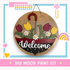 A wooden sign with a red umbrella handle and flowers, displaying the word "Welcome" in white cursive text. The sign is hanging on a wall, surrounded by various colorful stickers and illustrations.