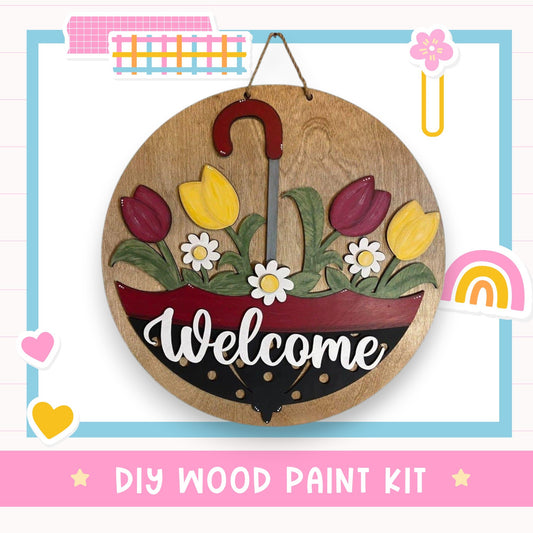 A wooden sign with a red umbrella handle and flowers, displaying the word "Welcome" in white cursive text. The sign is hanging on a wall, surrounded by various colorful stickers and illustrations.